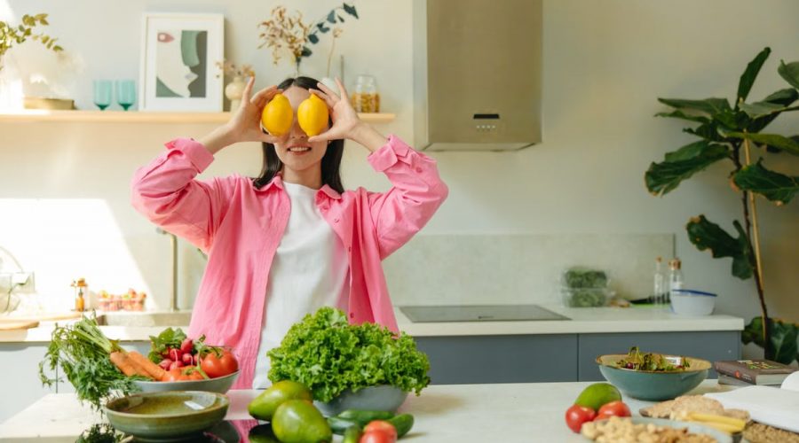 A woman playfully holding lemons over her eyes in a bright, lively kitchen setting filled with fresh vegetables.